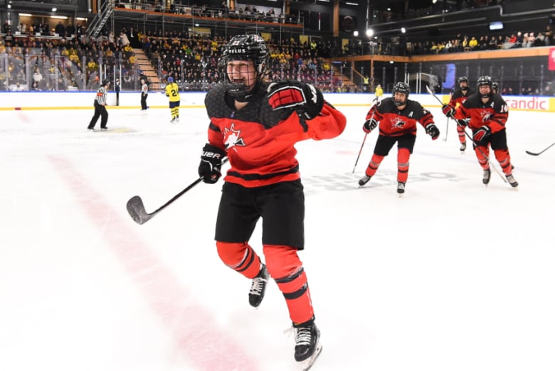 A female ice hockey player smiles and pumps her fist while celebrating on the ice with teammates.