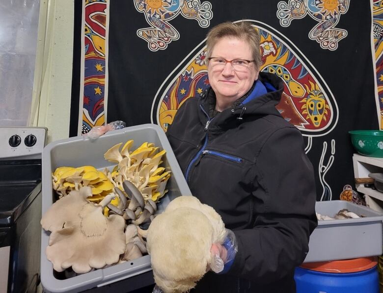 A middle-aged woman with short hair and glasses holds a container full of large edible mushrooms. Some are yellow and some are white. 