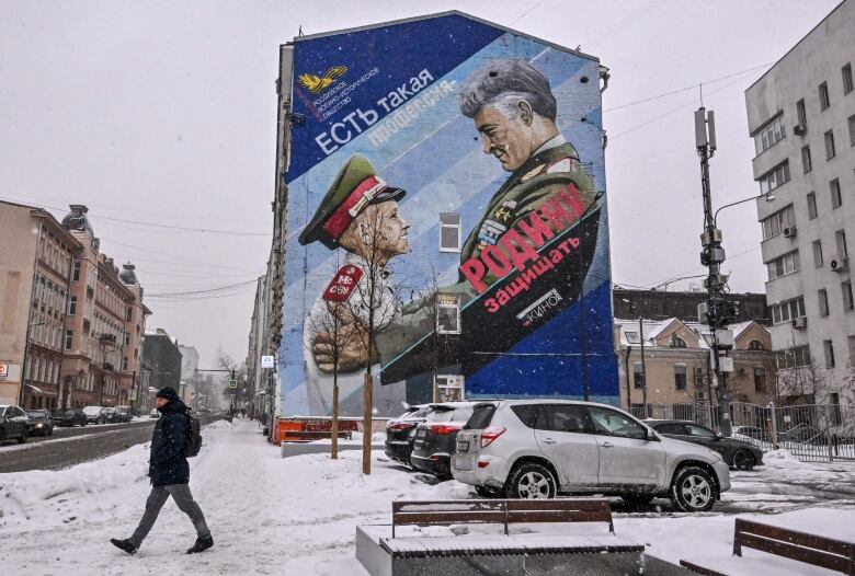 A man walks through on snowy ground in a parking lot with a mural on the side of a building shown in the background.