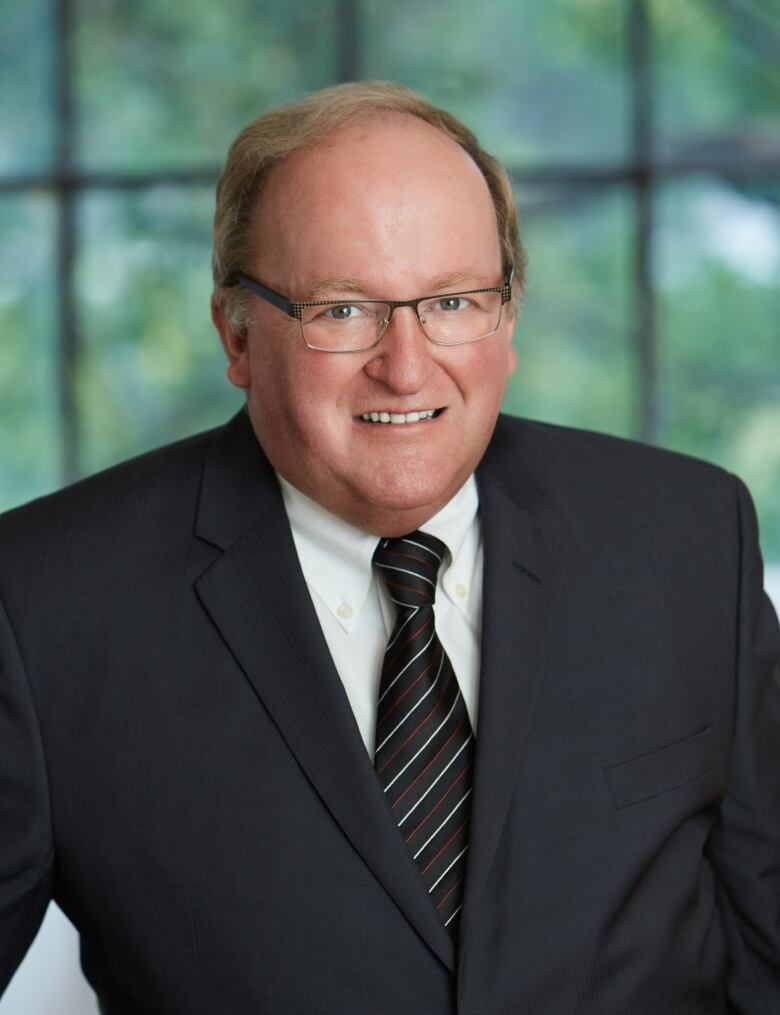 A bald spectacled man clad in a suit stands smiling.