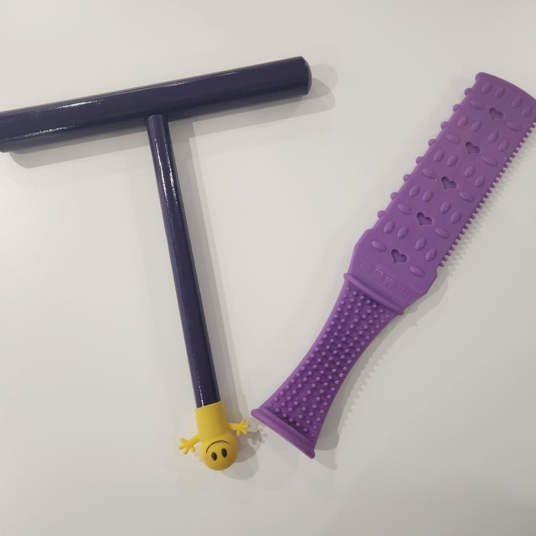 The device on the left is in a T shape, and has a small rubber stopper on the end with a yellow happy face. The device on the right is a purple rubber paddle with small ridges on it.