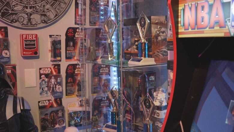 Action figures, still in their boxes, are stuck to a wall. A glass showcase displays multiple trophies.