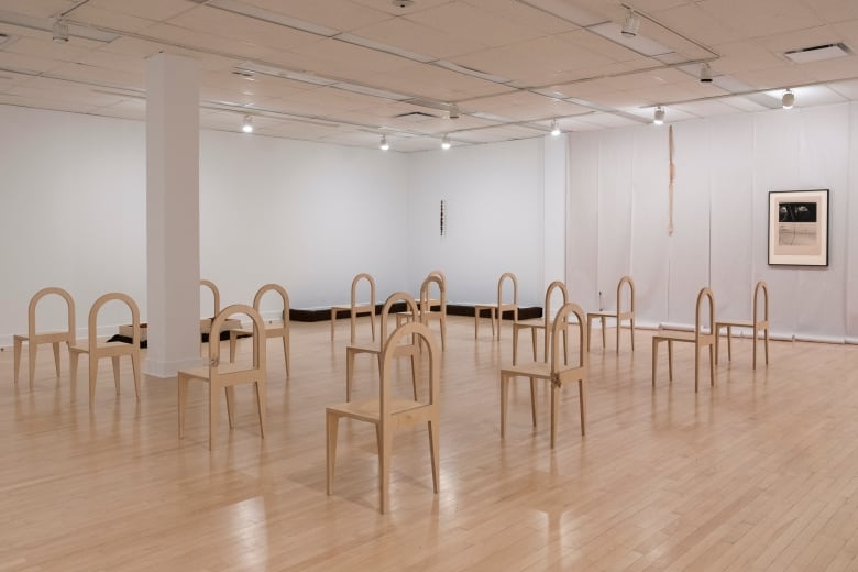 Framed artwork in the background on a wall made up of rolls of white paper, with chairs in a grid facing to the left in the foreground.