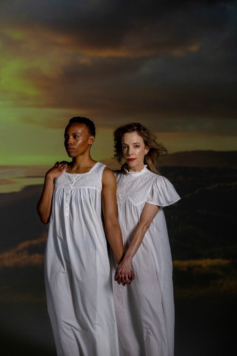 Actresses Amaka Umeh and Deborah Hay hold hands as they stand in white gowns in front of a backdrop of mountains and a sunset in a promotional image from Fall On Your Knees.