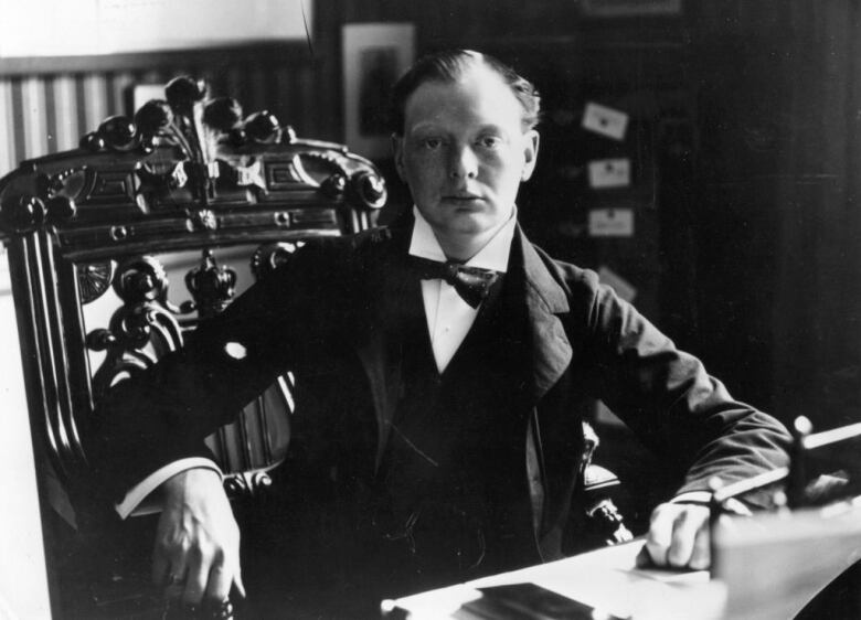 A young Winston Churchill sits at a desk wearing a bow tie and suit with his hair brushed back. His chair is wooden with ornate carvings and his desk has papers on them.