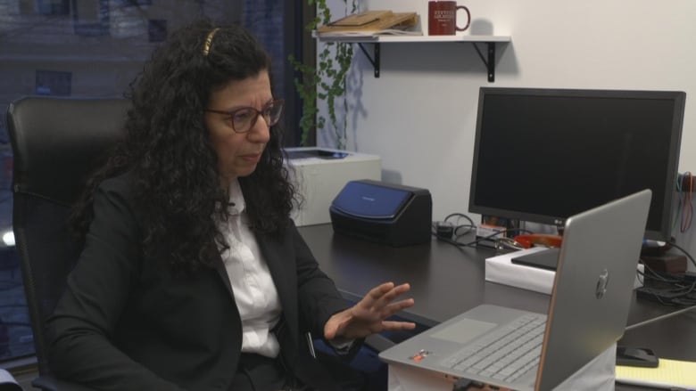 A woman with dark curly hair, wearing a white button-down shirt and a black blazer, sits at a desk in front of a laptop. She gestures with her hand as she talks to others on screen.