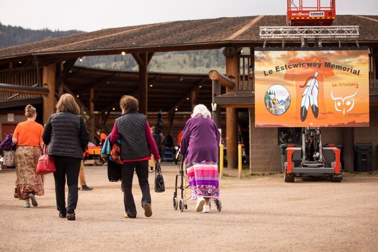 A group of people walk towards an amphitheatre, with an orange flag to the right memorializing those lost to Indian residential schools.