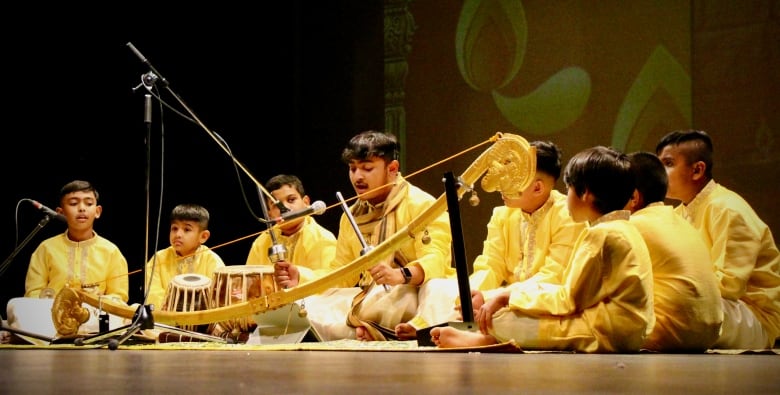 Children dressed in gold South Asian outfits sit on the floor singing and playing a Villu, or bow strung with bells.