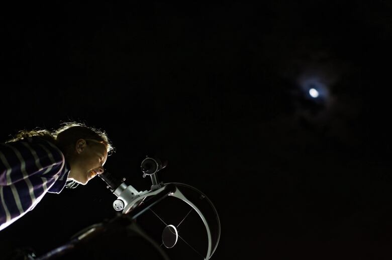 The photo shows a girl wearing a striped shirt looking through a telescope a the moon against a jet black sky. 