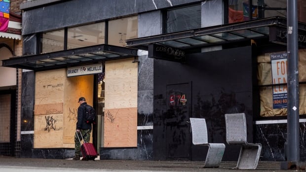 A man dragging a suitcase walks by boarded up shops