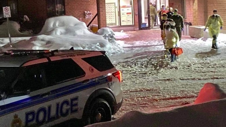 Emergency crews leave an apartment building at night. There's snow on the ground and a police vehicle outside.