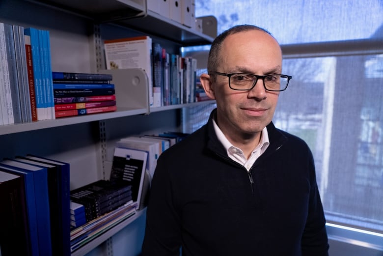 A man in glasses, a white button down shirt and black sweater stands in his office, with shelves filled with books and a window seen behind him.