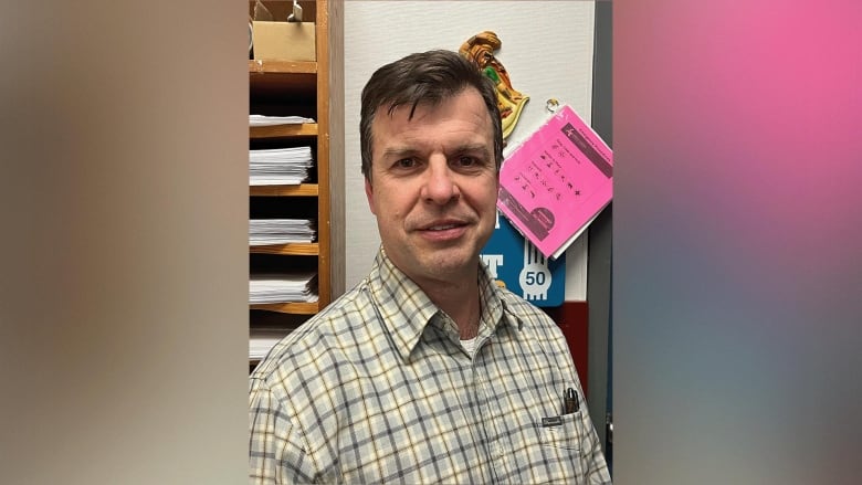 A man in a grey-and-yellow patterned shirt stands in a school office room. Flyers affixed to the wall and neat piles of papers are seen stacked on shelves behind him.