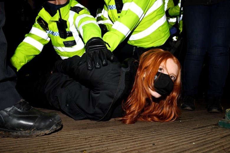 A red-headed woman wearing a face mask to protect against COVID-19 is held on the ground with her hands behind her back by multiple uniformed police officers.