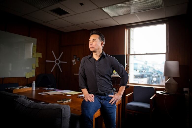 A man in blue jeans and a black shirt perches on the edge of the desk in an office with a window behind him.