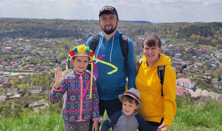 A family of four is posing for a group of photo on a pathway on a hill, that overlooks the entire community behind them.