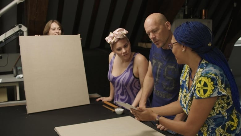 A woman with blue braided hair shows something to man and and a woman, as a third woman stands off to the side holding up a blank canvas.