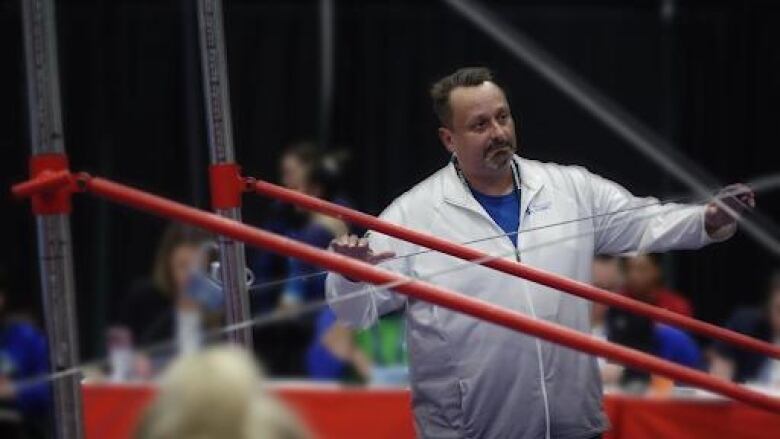 A man in a white jacket stands in an arena during a gymnastics competition.