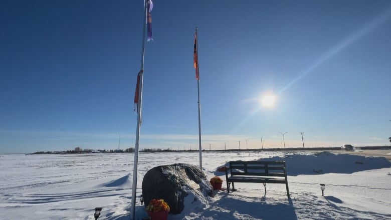 A boulder with a bronze plaque on it sits between two flag poles. It's surrounded by snow, under a blue sky and a shining sun.