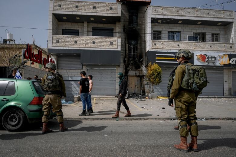 Two men in military uniform stand in the street near people in civilian clothing in front of a damaged building.