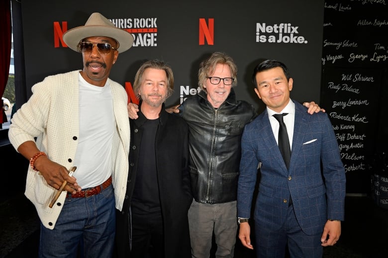 Four men stand shoulder-to-shoulder as they pose for a photo before a backdrop that says, "Netflix is a Joke".