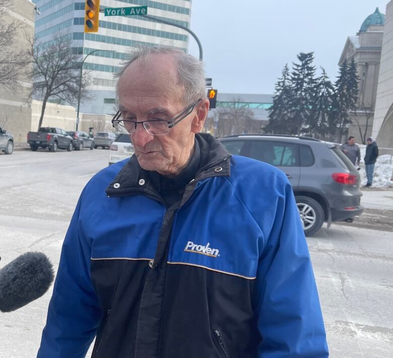 An old man with white, receding hair is wearing a blue and black jacket that says "Proven." He has eyeglasses and is looking at the ground.