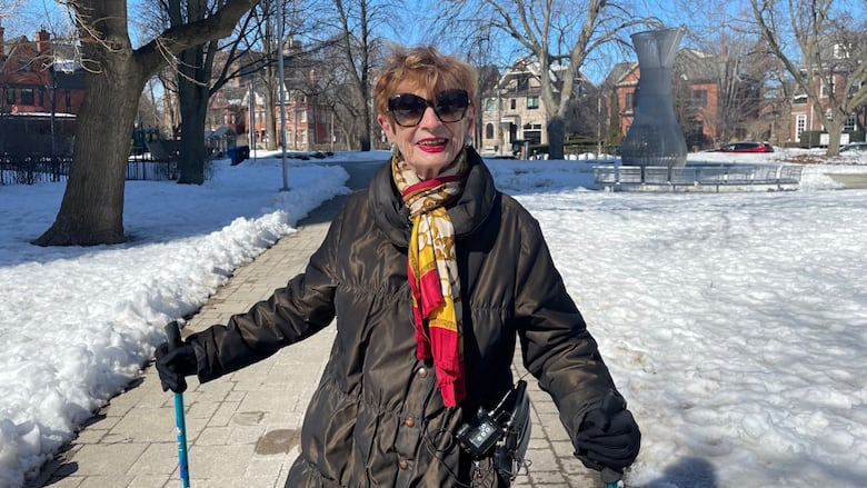 A 95-year-old woman stands in a park smiling at the camera, holding a walking stick in each hand.