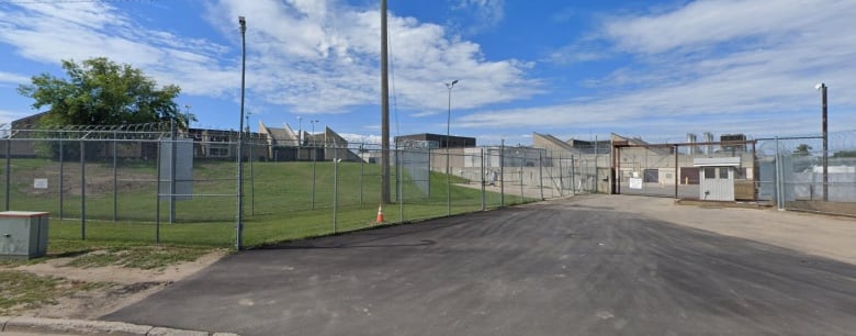 Barbed wire circles on top of the gated fence, which encloses a vast compound. With many large, concrete buildings.