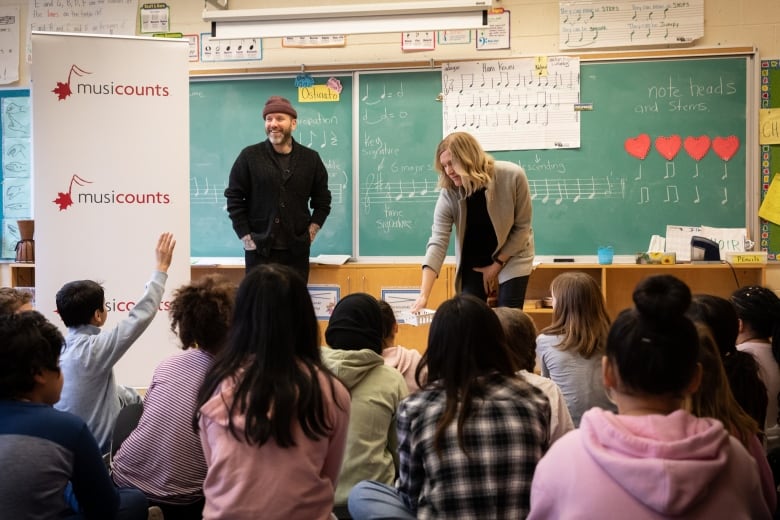 A woman bends to hand items to students seen seated, from behind. A man smiles at a student with his hand raised and music notes appear on the blackboard behind them.