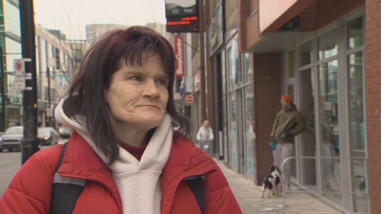 A woman with shoulder-length brown hair in a red jacket and white hoodie stands on a busy Halifax sidewalk.