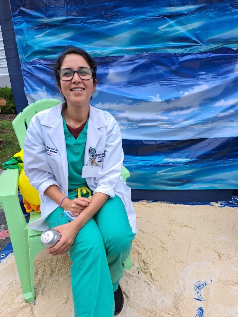 A smiling woman in hospital scrubs and a doctor’s coat sits in a patio chair.