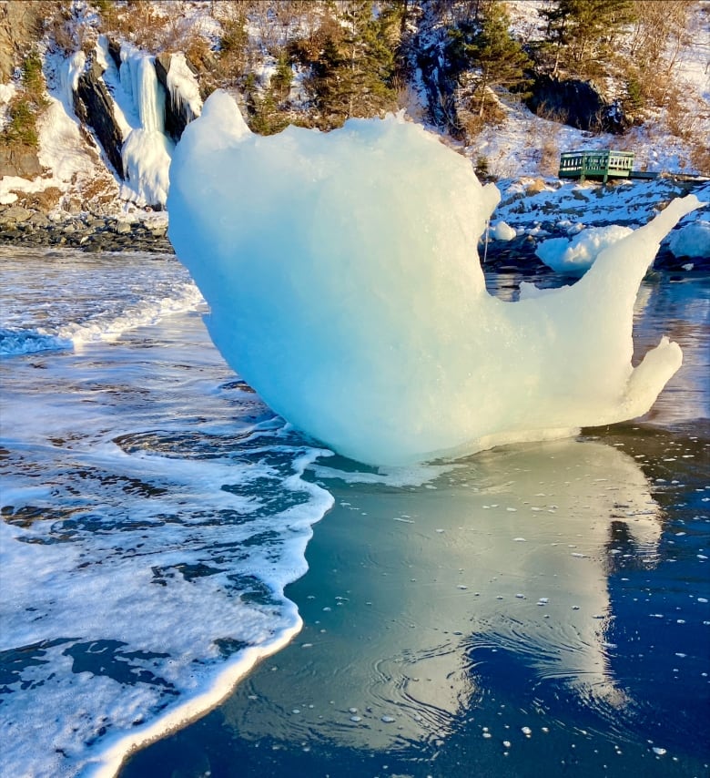 A large chunk of ice is reflected back by icy water with snowy cliffs in the background.