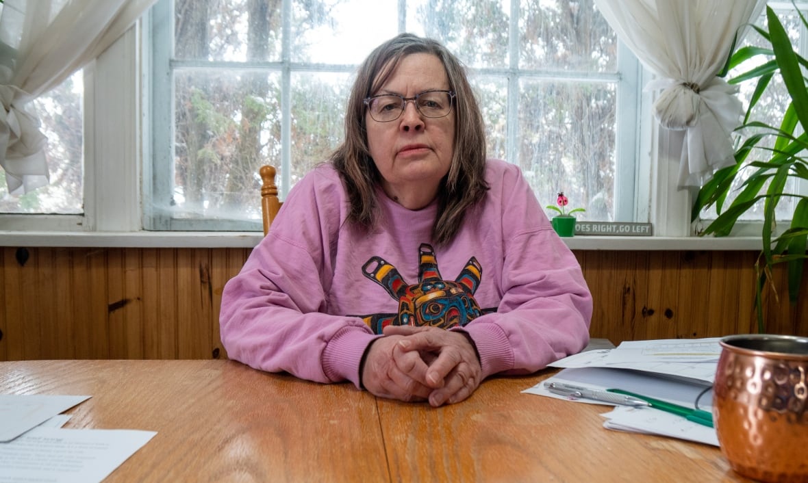 A woman with glasses and a pink sweatshirt sits behind a wooden table.