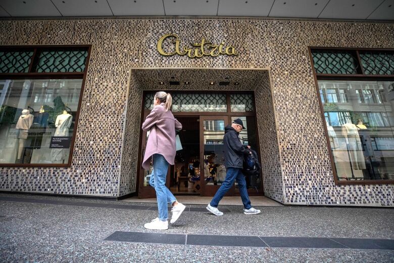 Two people wearing spring clothing walk in opposite directions past a storefront in downtown Vancouver. The word 