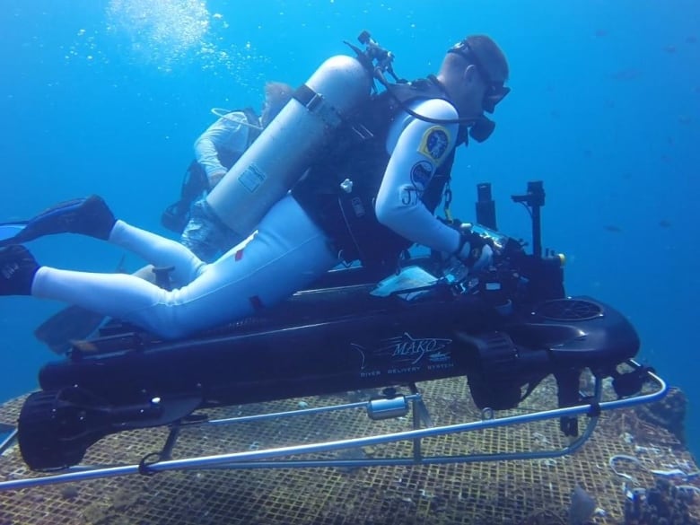 De Canadese astronaut Jeremy Hansen in een grijs duikpak zit in de oceaan bovenop een klein zwart vaartuig dat hem door het water voortstuwt.