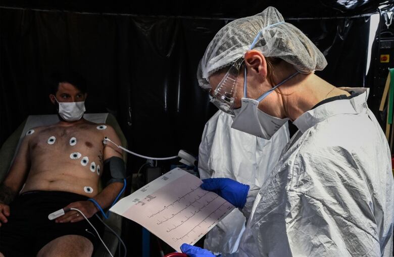 A man with electrodes still placed on him lies on a stretcher as a doctor reads his ECG readout with vertical spikes on the paper. 