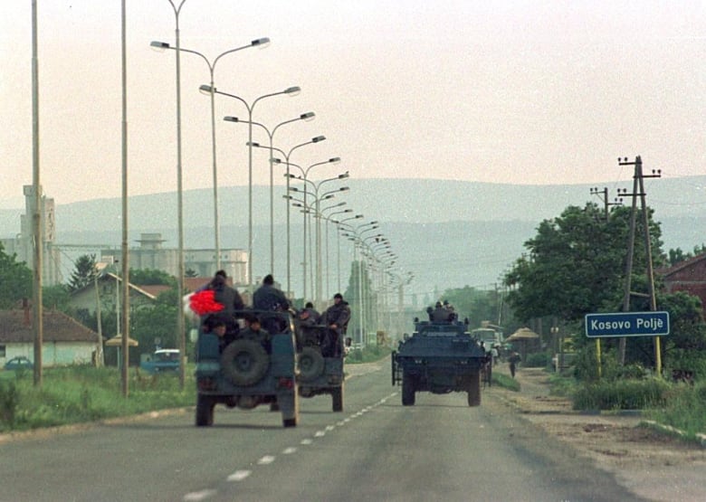 Tanks and military vehicles driving down a road in the direction of a hilly area in the distant background.