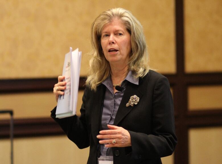 A woman speaks to an unseen audience at a conference, holding a report in one hand.  