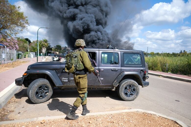 An armed soldier stands while looking in the direction of rising black smoke.