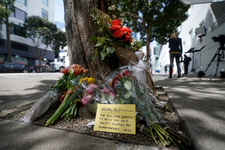 Flowers lie at the base of a tree, with a note that says BOB RIP - WE ARE ALL GOING TO MISS YOU. AND YOU WILL ALWAYS BE REMEMBERED. 