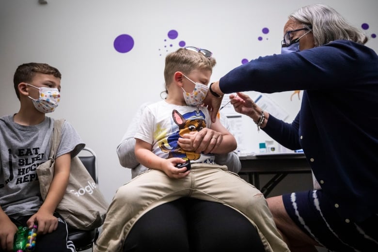 A nurse wearing a mask holds a syringe into a young boy's arm as he wears a mask and sits on his mother's lap, while another boy wearing a mask watches