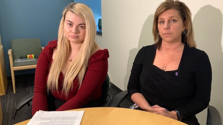 A woman with long blond hair wearing a red shirt sits at a table with another woman with shoulder-length brown hair and a black shirt. 