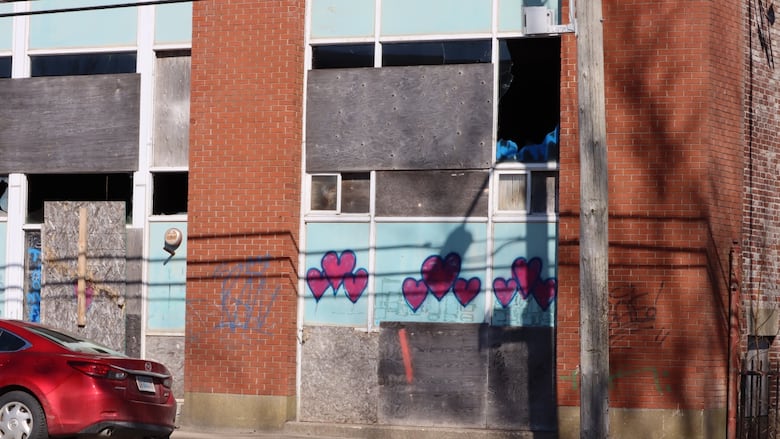 A derelict brick building with light blue panels under the windows is shown with boarded up and broken windows.