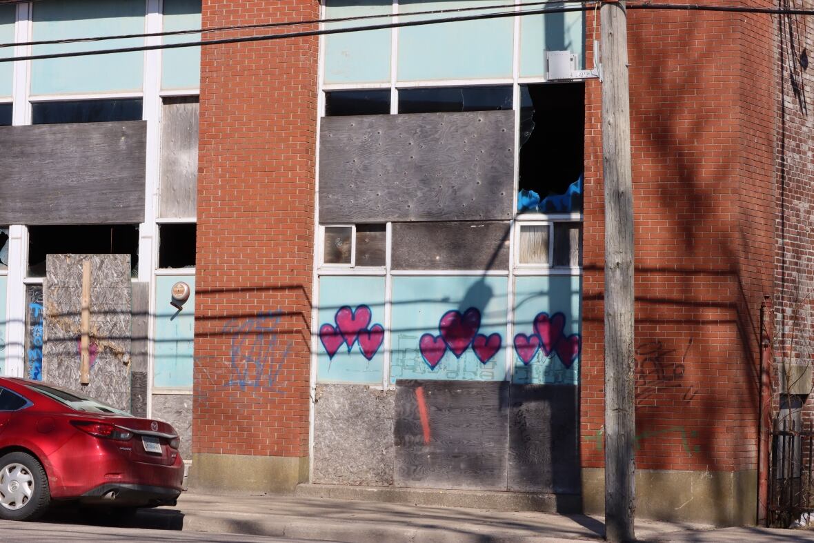 A derelict brick building with light blue panels under the windows is shown with boarded up and broken windows.