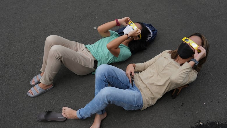 Two people are shown lying on pavement wearing special glasses and looking skyward.