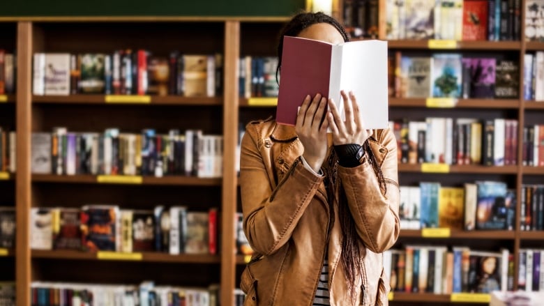 Person wearing a brown leather jacket stands in a book store, holding an open book closer to their face.