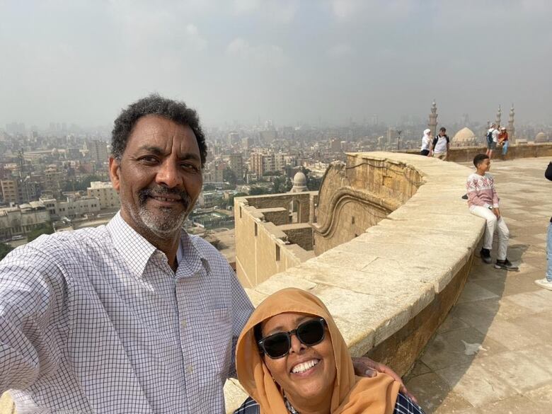 A man wearing a checkered shirt stands beside a woman wearing sunglasses and an orange shawl.