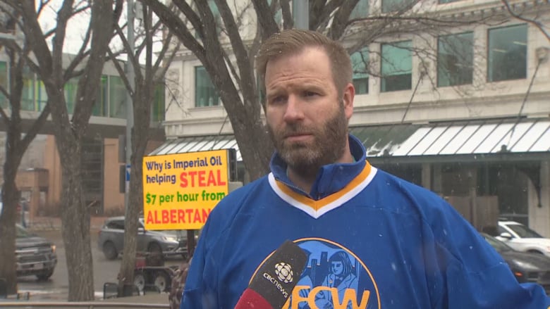 A man stands in front of a CBC News mic. In the background, a poster sign says "Why is Imperial Oil stealing $7 per hour from Albertans?"