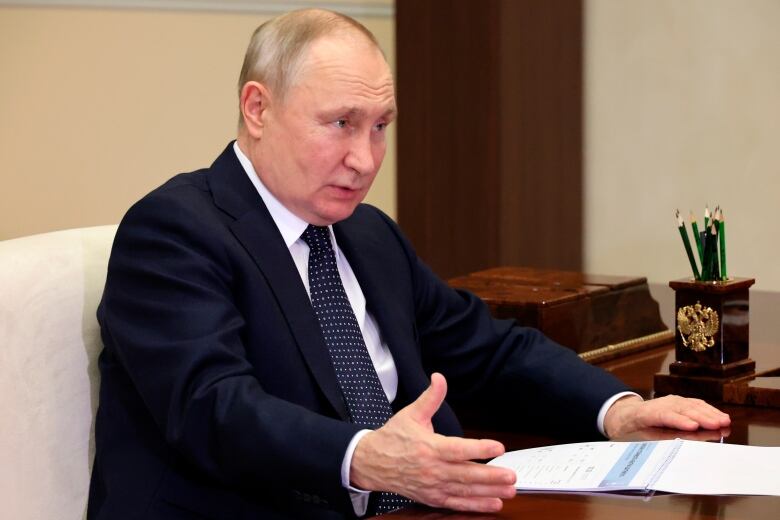 Russian President Vladimir Putin, wearing a dark suit and tie, talks with sitting at a desk.