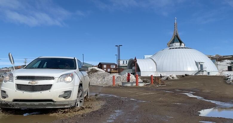 A white Chevrolet SUV splashes up water while driving through a large pothole on a wet, dirt road in Iqaluit, with Iqaluit's iconic Igloo church in the background.